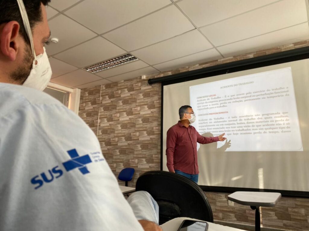 Em primeiro plano, a imagem mostra um colaborador do ISAC sentado, de perfil, assistindo uma palestra sobre acidente de trabalho. Em segundo plano, à frente do projetor, o técnico de segurança do trabalho da unidade faz a apresentação da palestra.