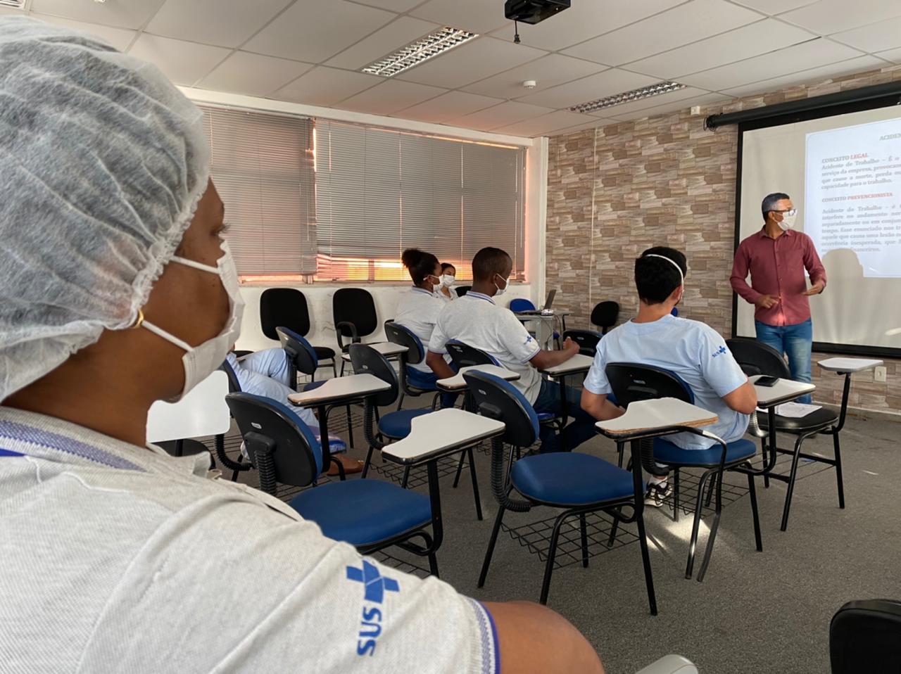 O técnico de segurança do trabalho está à frente do projetor de apresentação. Em primeiro plano, aparecem os membros da CIPA sentados nas cadeiras do auditório, de costas para a câmera e com os olhares direcionados para a tela de projeção.