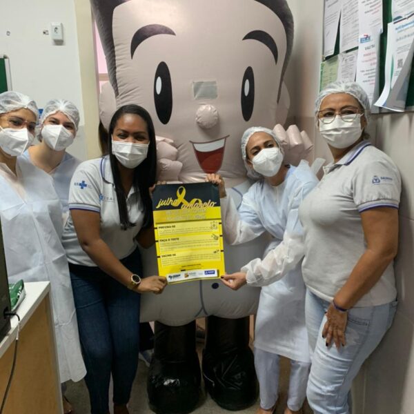 Colaboradoras da unidade posam para foto ao lado do Isaquinho, boneco que representa a marca ISAC. Todas estão de máscara, seguram um cartaz sobre o Julho Amarelo e sorriem para câmera.