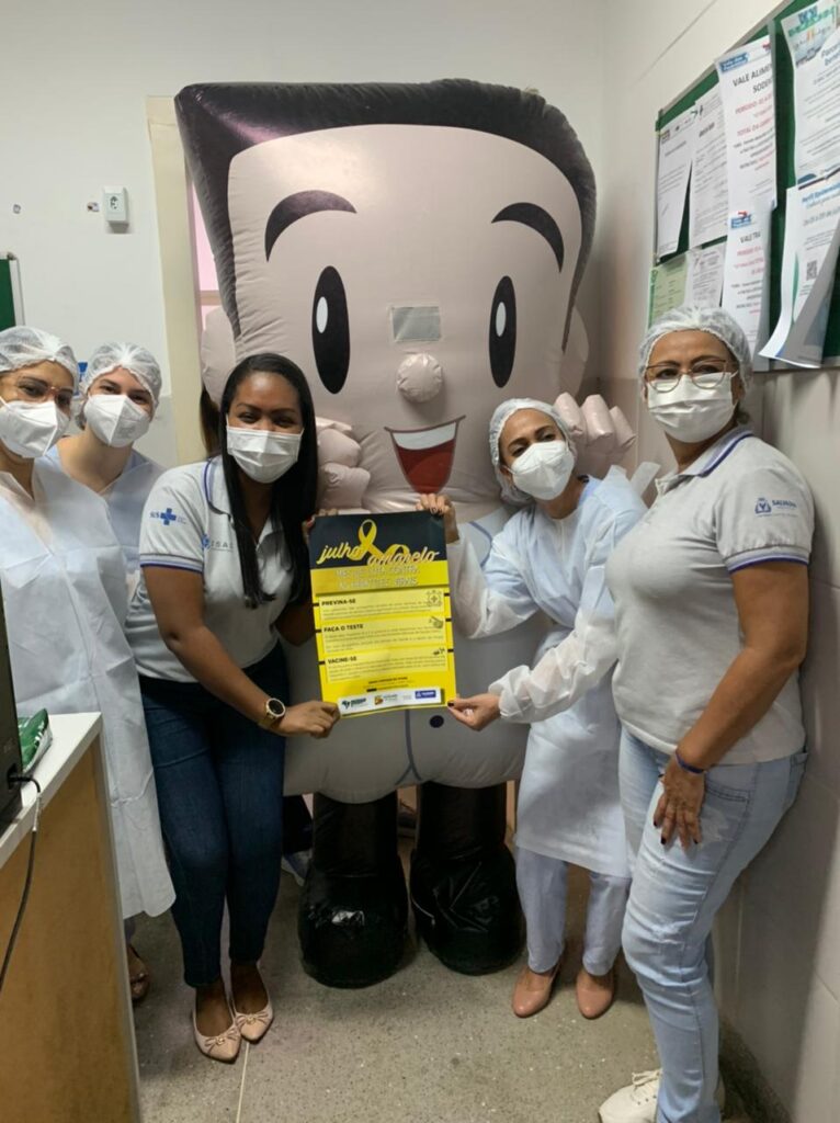 Colaboradoras da unidade posam para foto ao lado do Isaquinho, boneco que representa a marca ISAC. Todas estão de máscara, seguram um cartaz sobre o Julho Amarelo e sorriem para câmera.