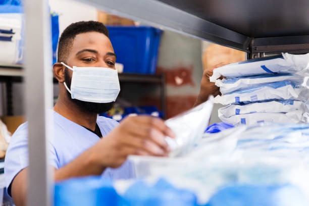 Em primeiro plano, aparece uma prateleira com insumos hospitalares com a imagem desfocada. Ao fundo, um homem com máscara e vestido com fardamento hospitalar arruma os materiais na prateleira.