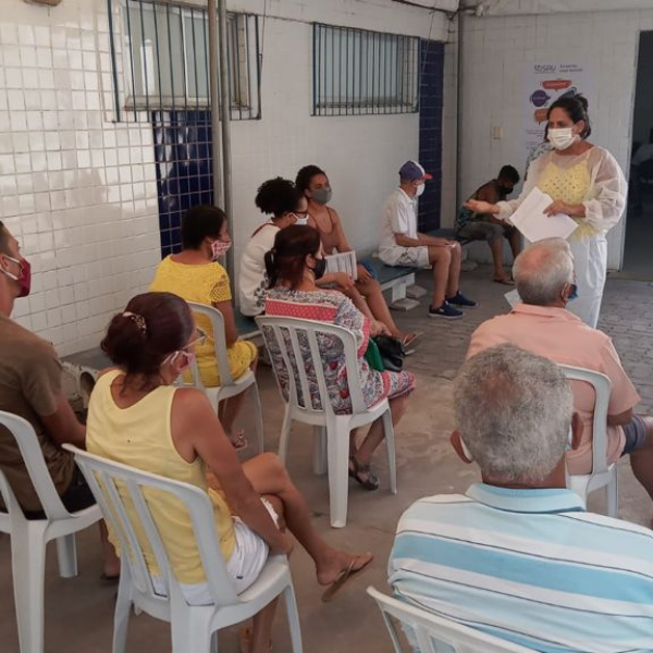 No primeiro plano, algumas pessoas de costas para câmera estão sentadas em cadeiras com o devido distanciamento uma das outras. No funfu, uma mulher de pé, usando máscara e avental, ministra a palestra.