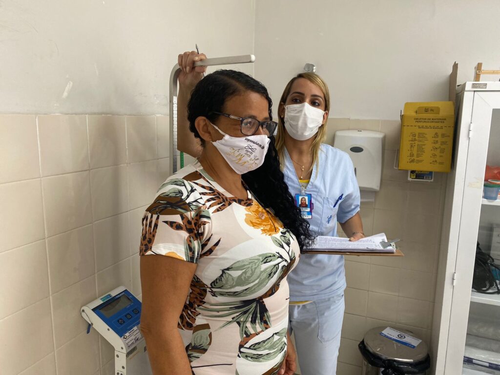 Mulher está de pé em uma balança, enquanto outra com fardamento azul realiza a aferição da altura. Ambas utilizam máscara.